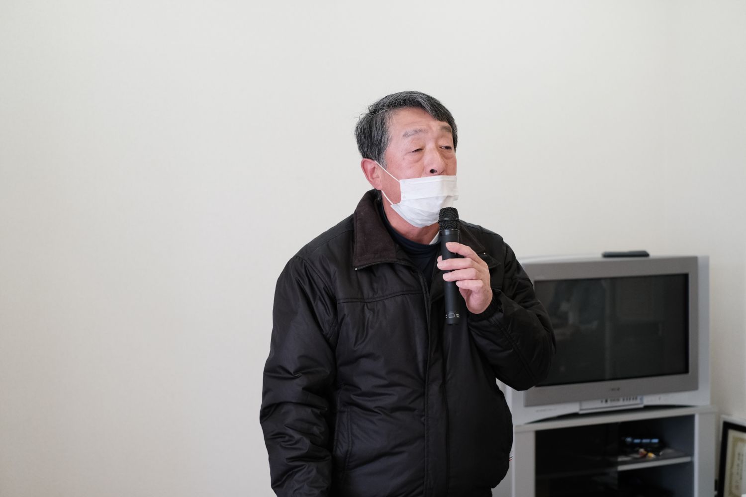 黒いジャンパーと白いマスクを着けた年配の男性が、マイクを持って話しをしている写真