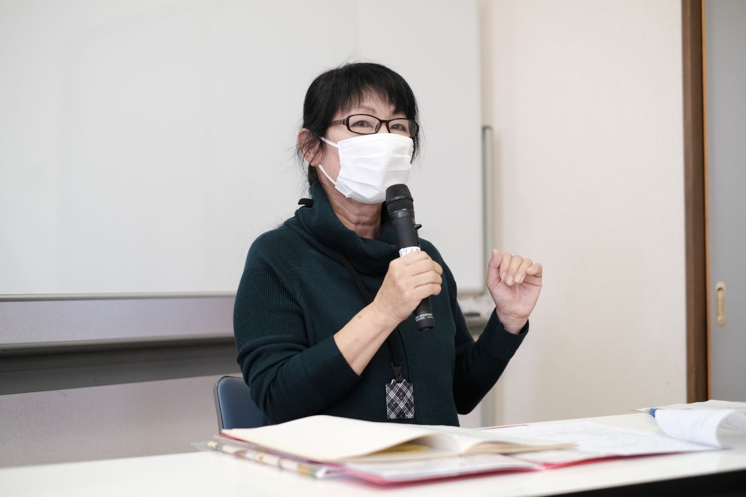 白いマスクを着けた黒縁メガネの女性が、開いた資料の前で黒いマイクを持ちながら話している写真