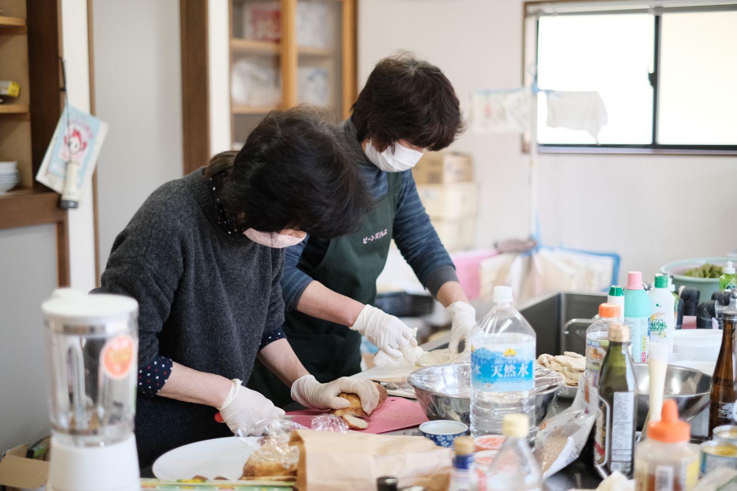 調理室のようなところで、ミキサーや複数の調味料が所狭しと並べられ手袋を着けた2人の女性が、パンを手に取りながら料理を行っている調理中の写真