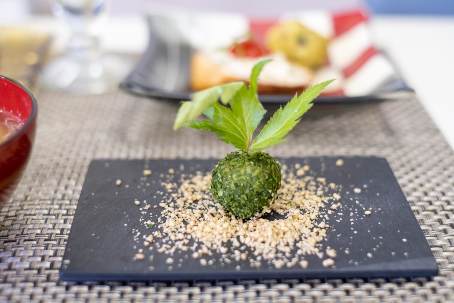 黒い皿の中央に、緑色のトウキの葉の粉で覆われた球状の料理の上にトウキの葉が飾られた写真