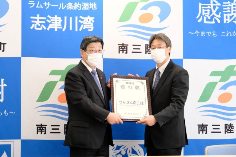 南三陸町のバックパネルの前で、額入りの登録証を一緒に持っている2人のスーツ姿の男性が並んでいる記念写真