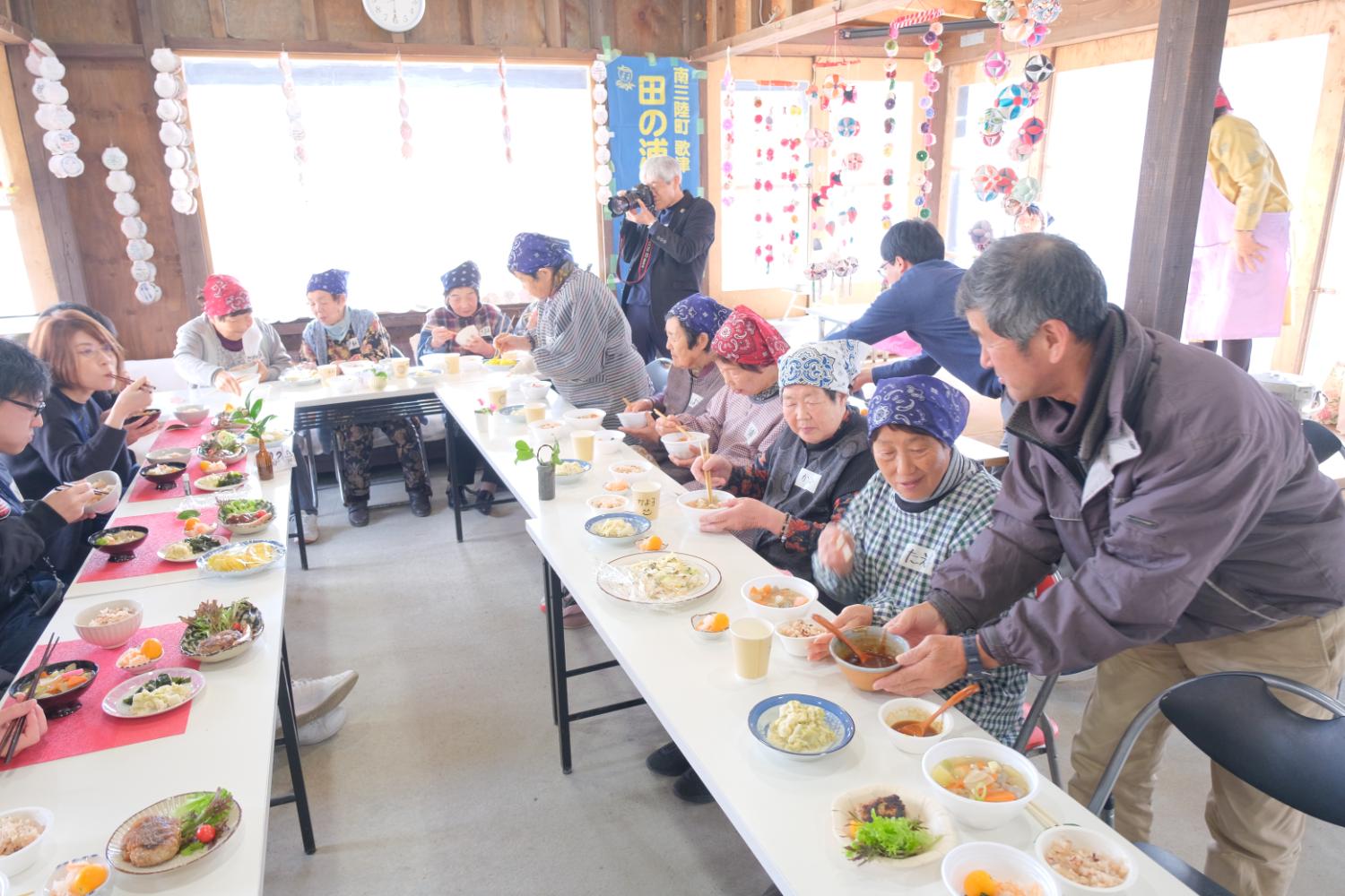 高齢の女性たちが中心となり、木造の明るい室内で長テーブルを囲み、手作りの料理を囲んで交流している様子の写真