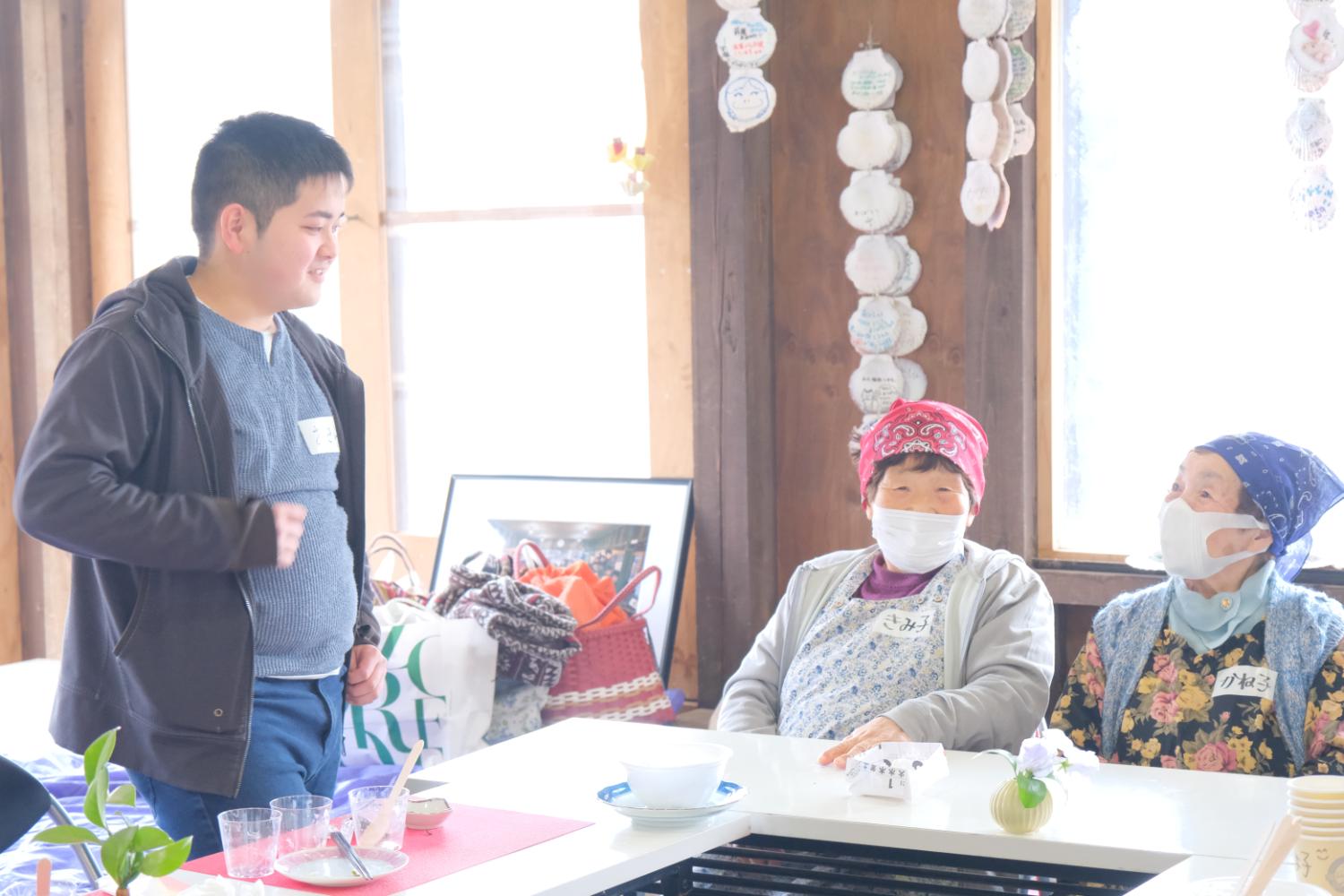 室内で、マスクを着けた赤いバンダナの女性と青いバンダナの女性が座っており、向かいにいる男子生徒と会話をしている様子の写真