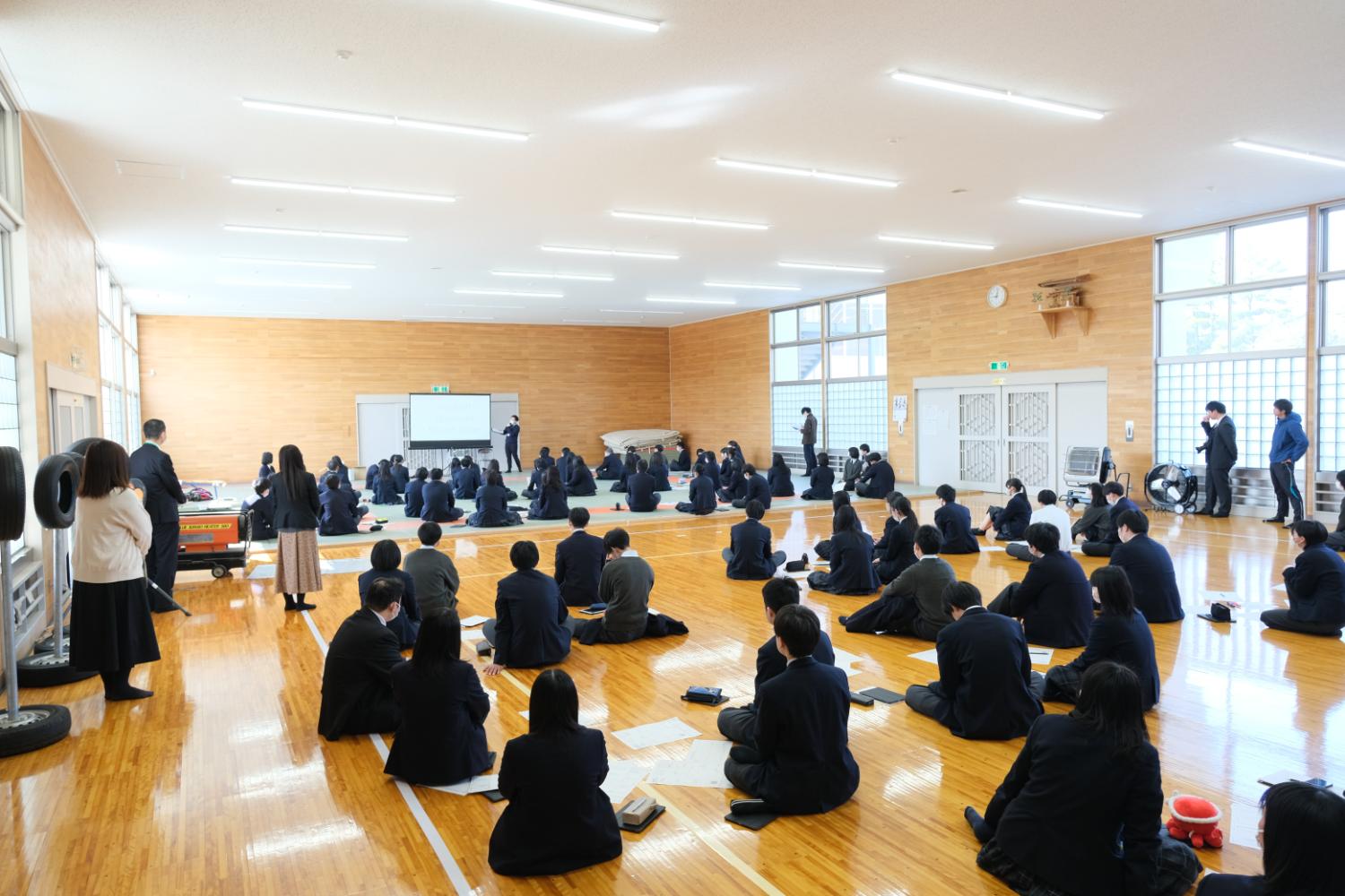 広い体育館のような木製の床の部屋で、多くの生徒が床に座って前方のスクリーンを見ている様子が写っている写真
