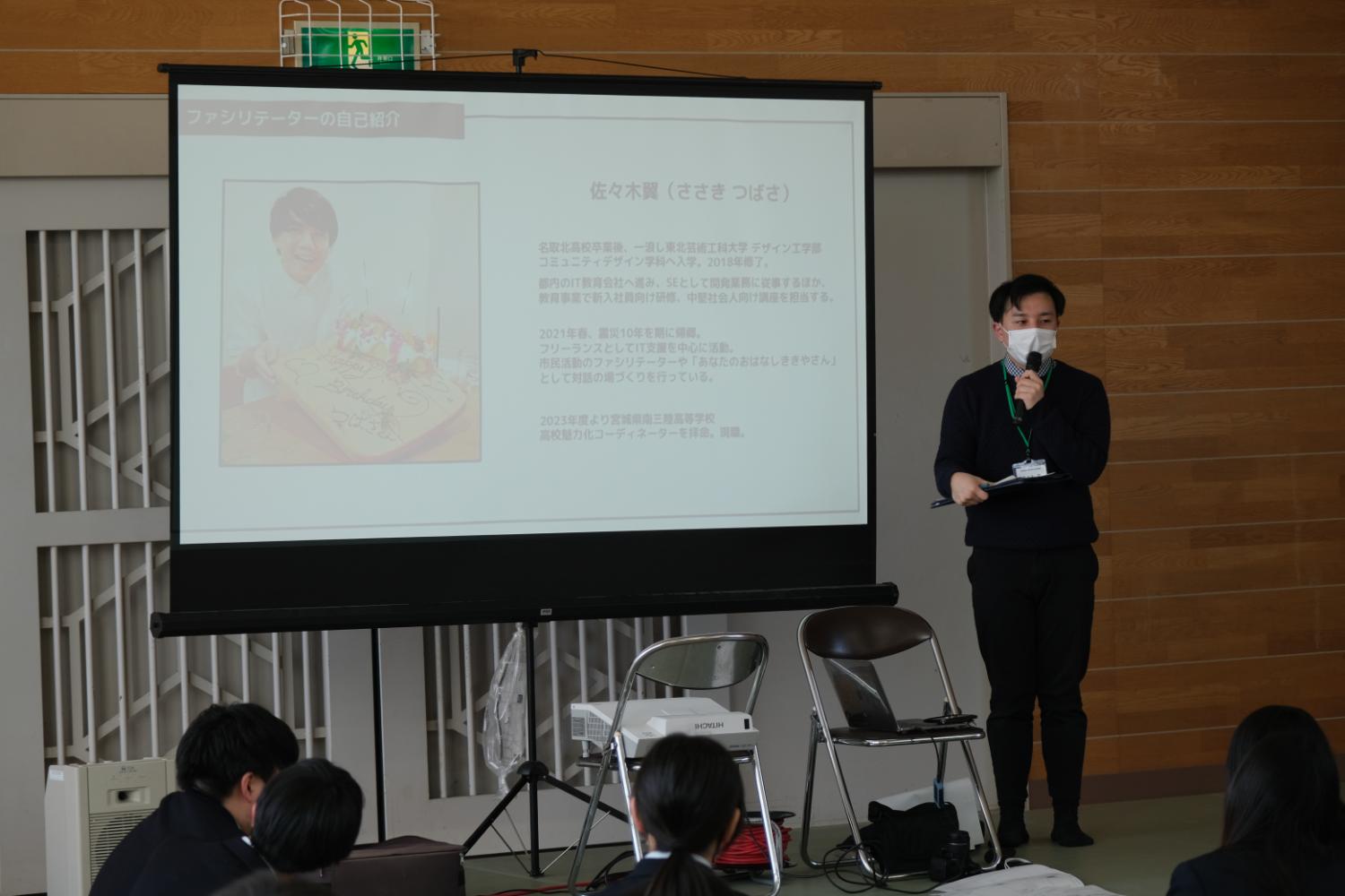 木製の壁の前に設置されたスクリーンにスライドが映し出されており、スクリーンの右側で名札をつけた男性がマイクを持って説明している様子が写っている写真