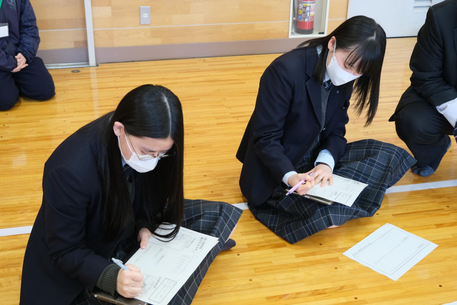 制服姿の女子生徒2人が床に座り、用紙に記入している様子の写真