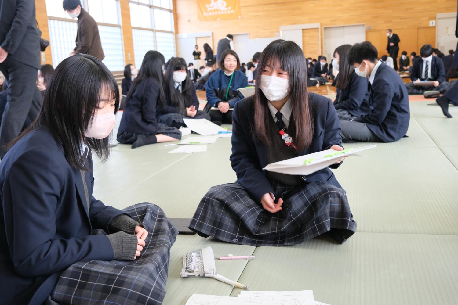 制服を着た生徒たちがグループごとに座り、ノートやプリントを見ながら意見を交わしている写真