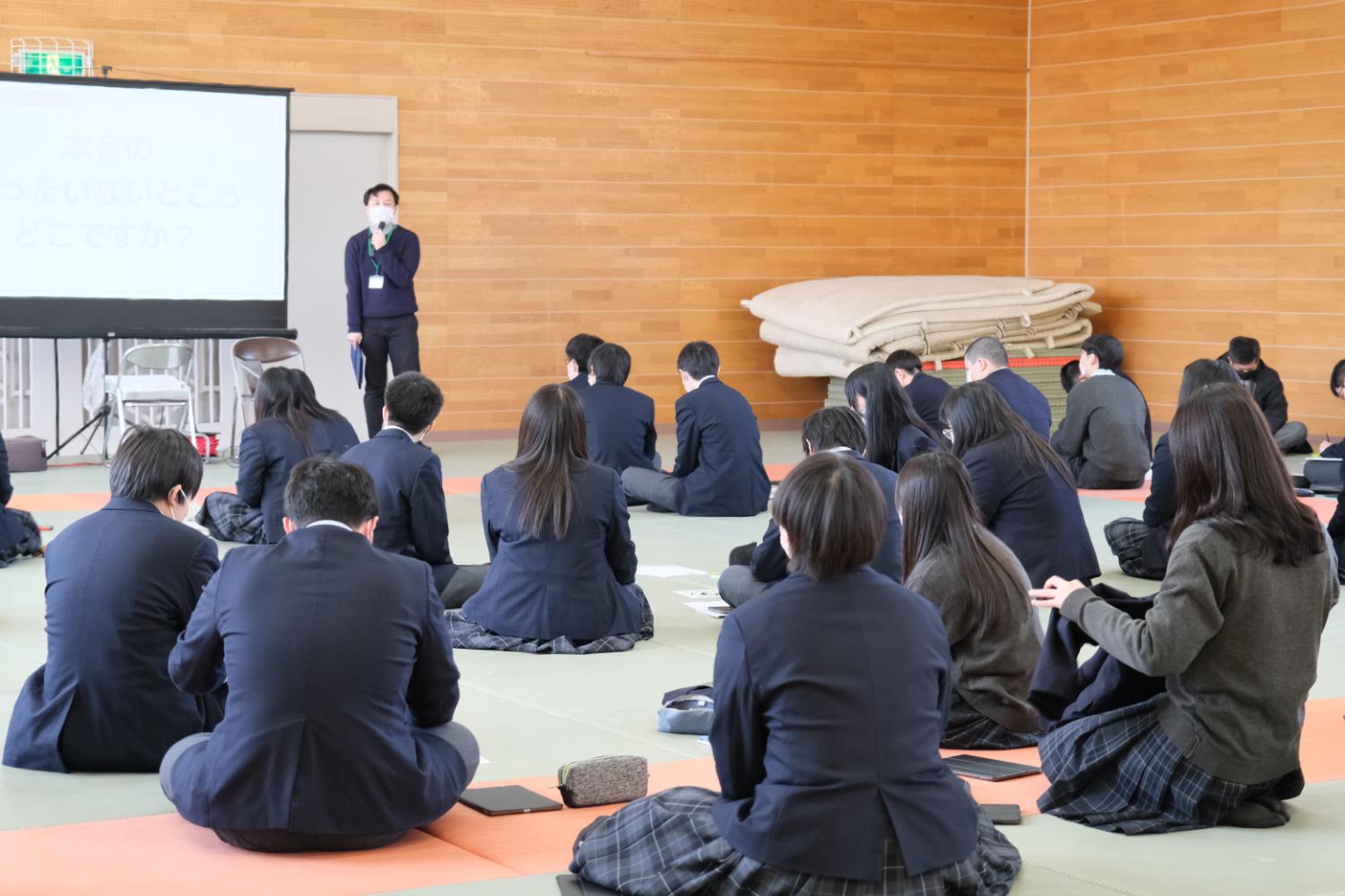 体育館のような広い部屋で、生徒たちが床に座って前方のスクリーンとマイクを持った男性の説明を聞いている様子が写っている写真