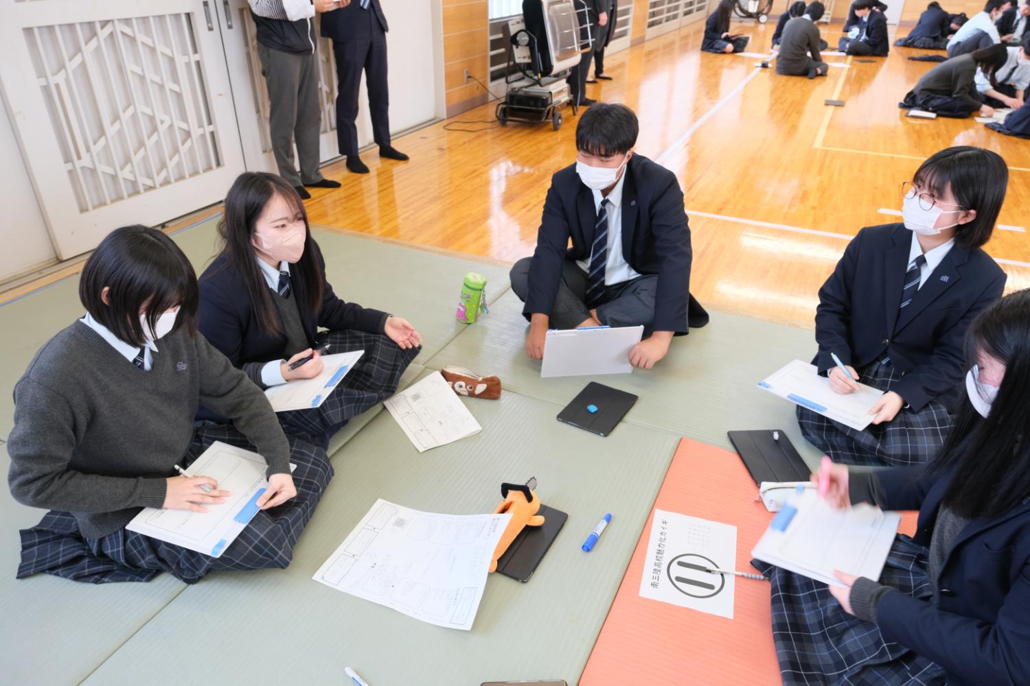 制服を着た五人が床に座り、ノートや資料を見ながら話し合っている様子が写っている写真