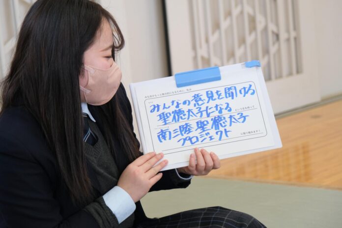 制服を着た女子高校生が、白いボードに「みんなの意見を聞いて聖徳太子になる南陵聖徳太子プロジェクト」と青い文字で書かれたものを持っている写真