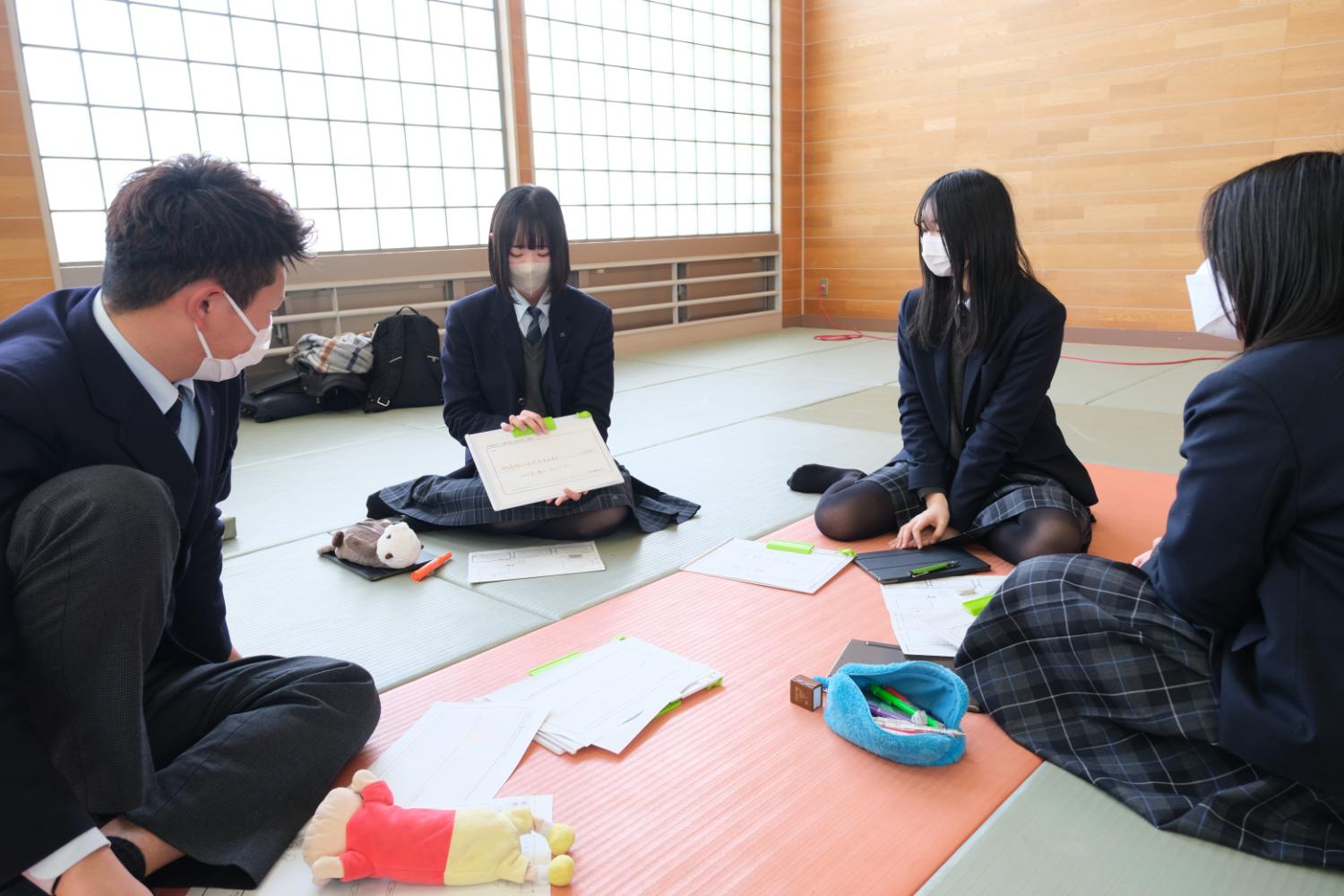 広い部屋の畳の上で、制服を着た四人が座り、ノートや資料を見ながら話し合っている様子が写っている写真