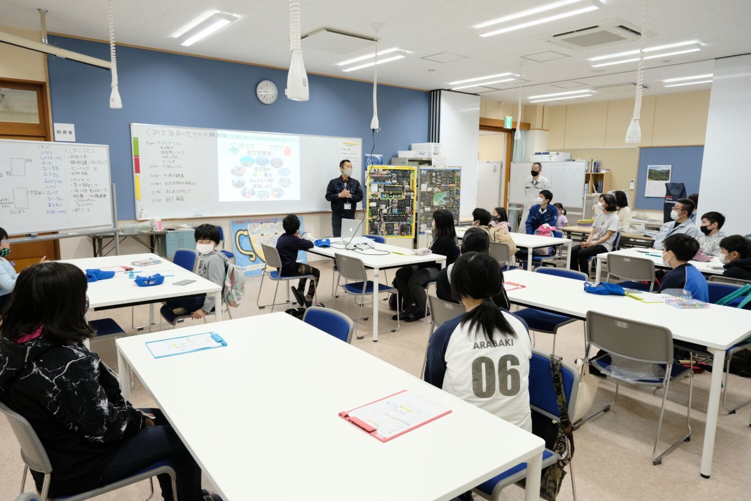 施設内の前方のホワイトボードの右側でマイクを持ち説明をしている男性の前で参加者が話を聞いている写真