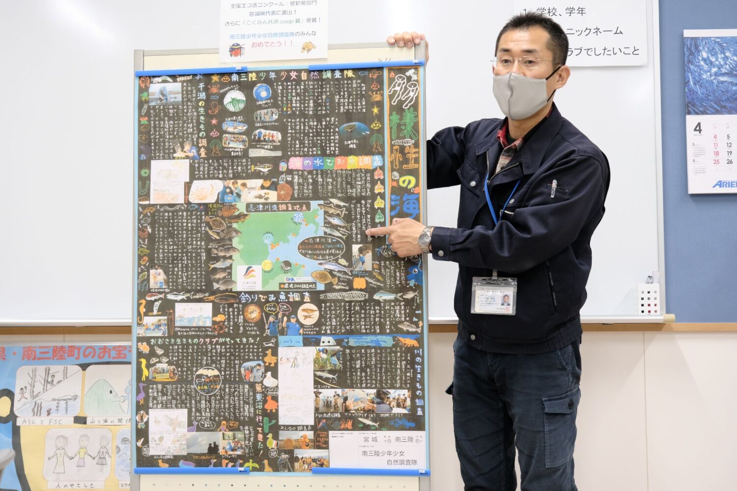 屋内で一人の男性がマスクを着用して立ち、大きなカラフルな壁新聞を指差しながら説明し、壁新聞には手書きの文字やイラスト、地図、写真などが所狭しと並んでいる写真