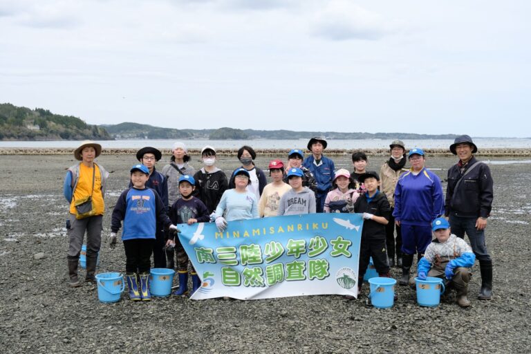 岩場の海岸で青いバケツを前に並べて「南三陸少年少女自然調査隊」と書かれた横断幕を掲げ、参加者の大人や子供たちが並んでいる様子の写真
