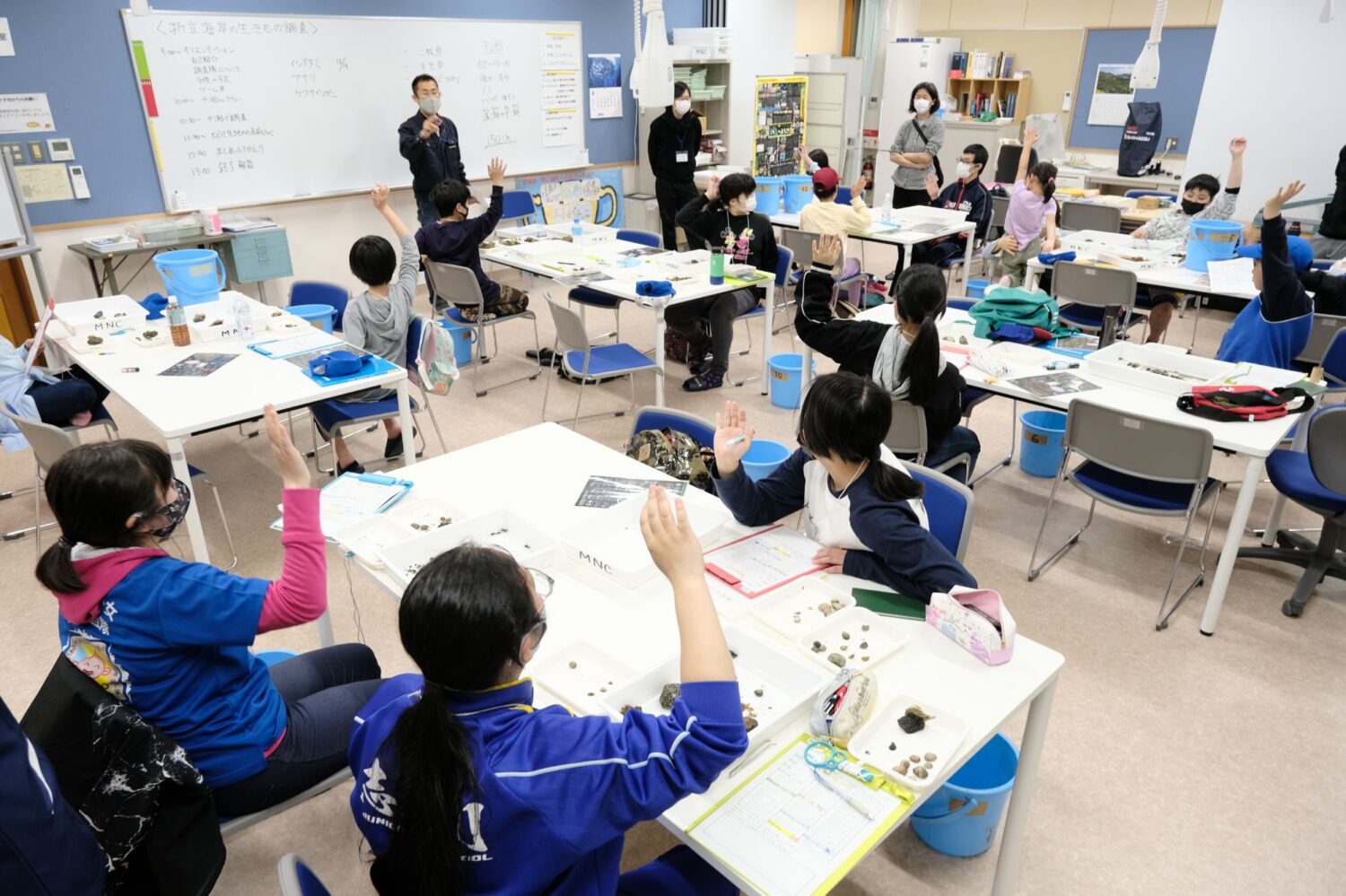 教室内でたくさんの子どもたちが座り、前方のホワイトボードの前に立った男性の話に手を挙げている写真