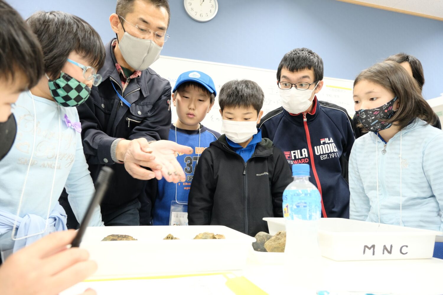 男性が両手で小さな生き物を見せながら、マスクをつけた子どもたちが男性を囲んで熱心に観察している様子の写真