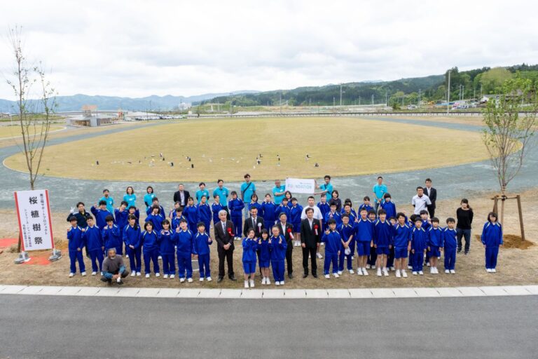 「桜植樹祭」と書かれた看板のそばで、青いジャージを着た生徒たちとフォーマルな服装の大人たちが広い陸上競技場の前に並んでいる様子を写した写真