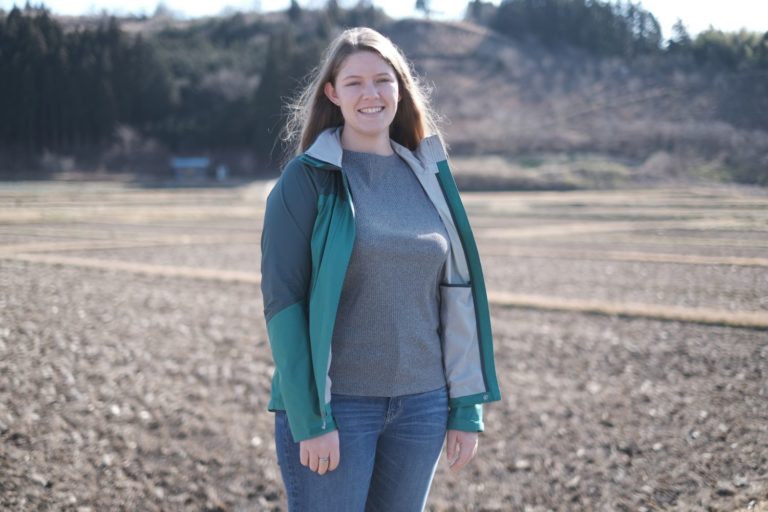 何も植えられていない畑の真ん中に笑顔で立つ、緑色のジャケットを着たケーリー・ワードさんをアップで撮影した写真