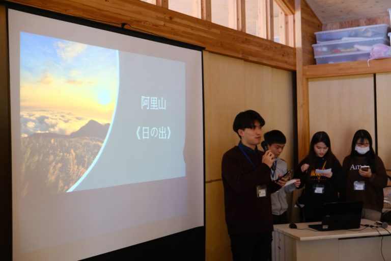 「阿里山(日の出)」と書かれた文字と雲が望める高台から朝日を撮影した写真が映し出されたスクリーンが設置され、右側に4人の学生が並び、左側の男子学生が右手にマイクを持ち話をしている様子の写真