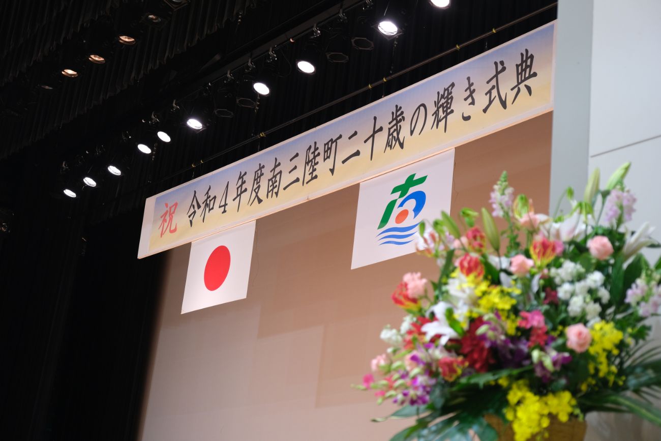 成人式会場のステージ上部に掲げられた「祝 令和4年度南三陸町二十歳の輝き式典」の横断幕と日本国旗、町章が写っている写真