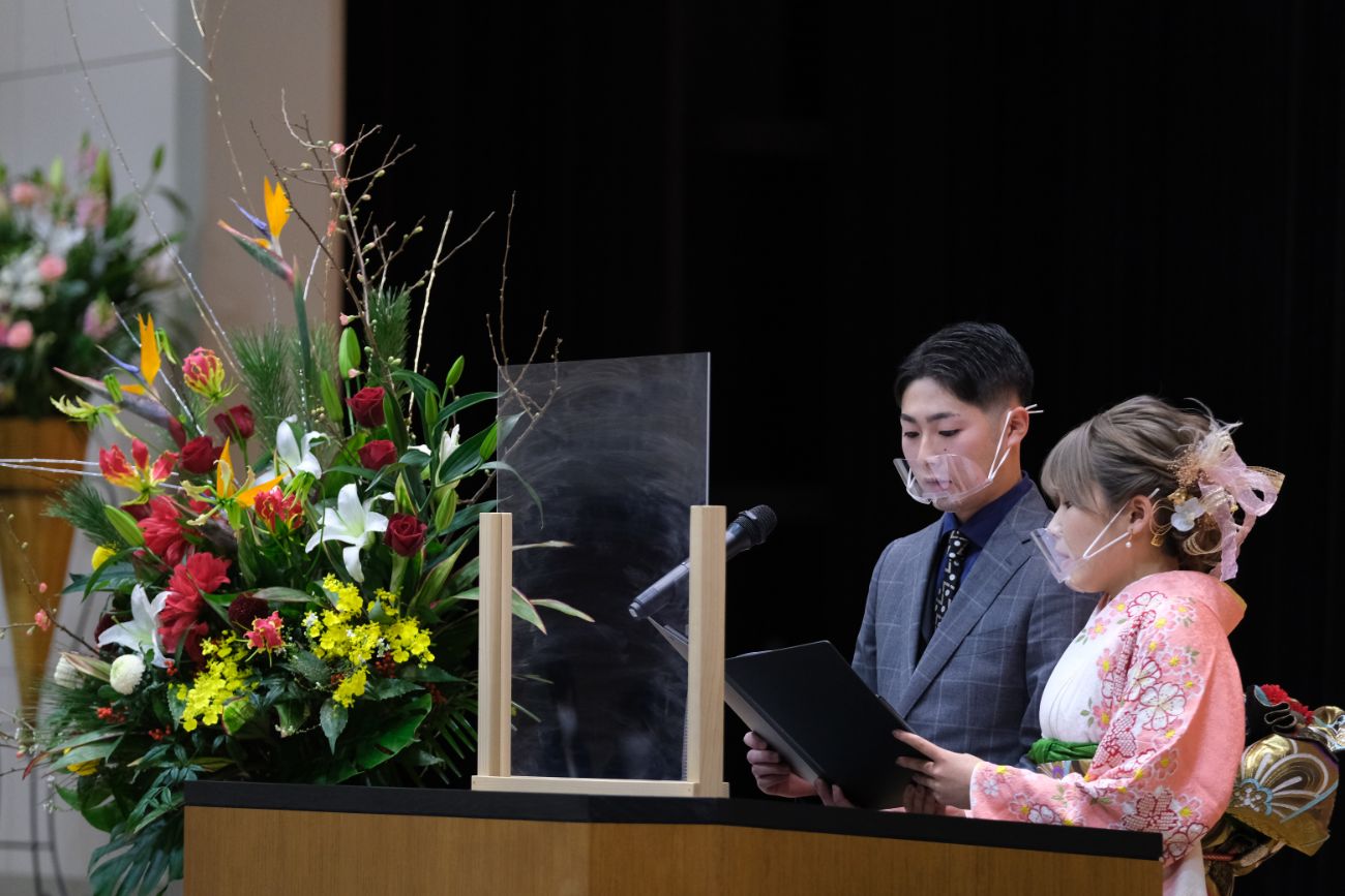 演台の前に立つ新成人の和装とスーツ姿の男女2人がそれぞれマイクに向かって原稿を読み上げている様子の写真