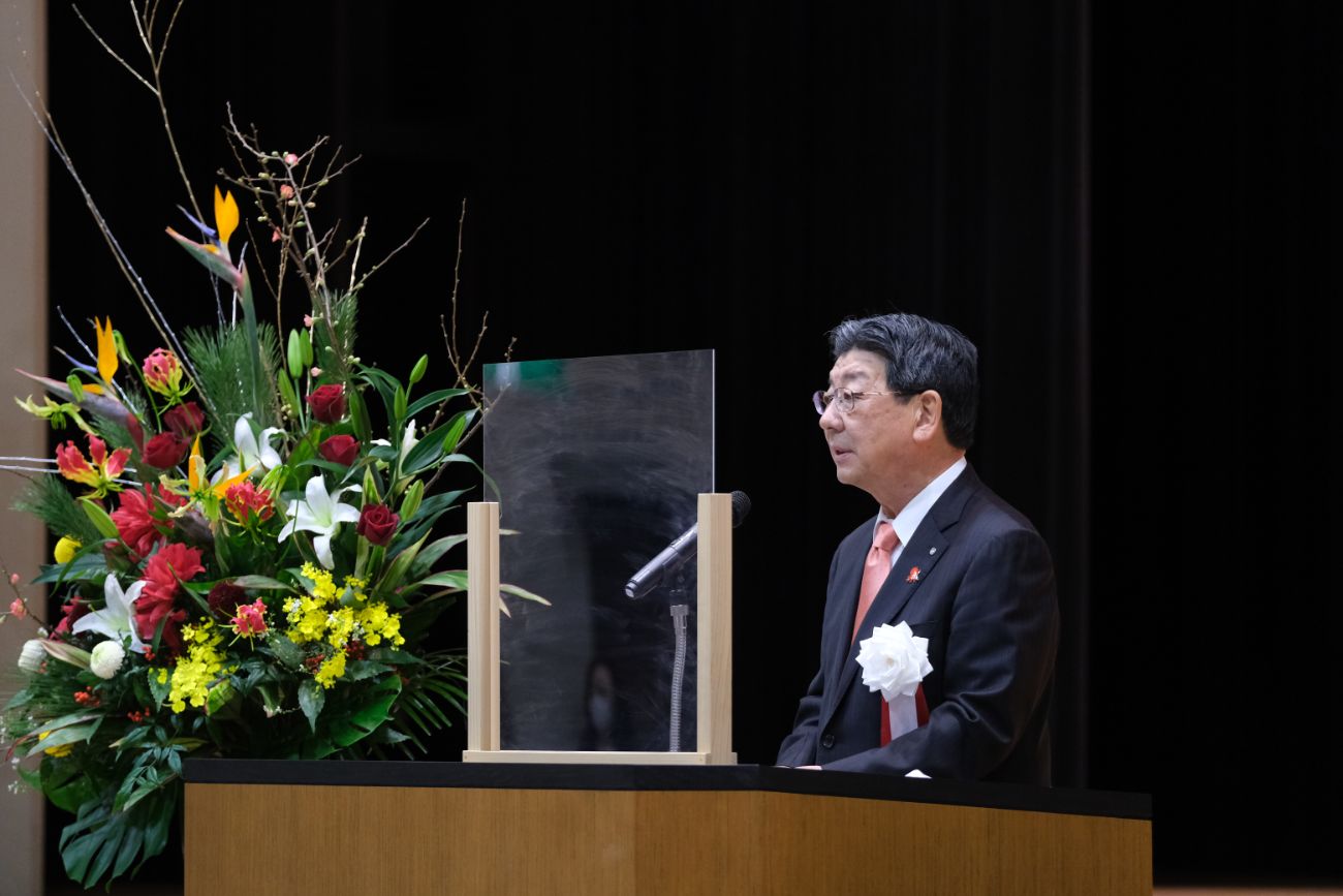 色とりどりの生花が飾られ、飛沫防止のアクリル板が設置された壇上でスーツ姿の佐藤仁町長が演台に立って挨拶している写真
