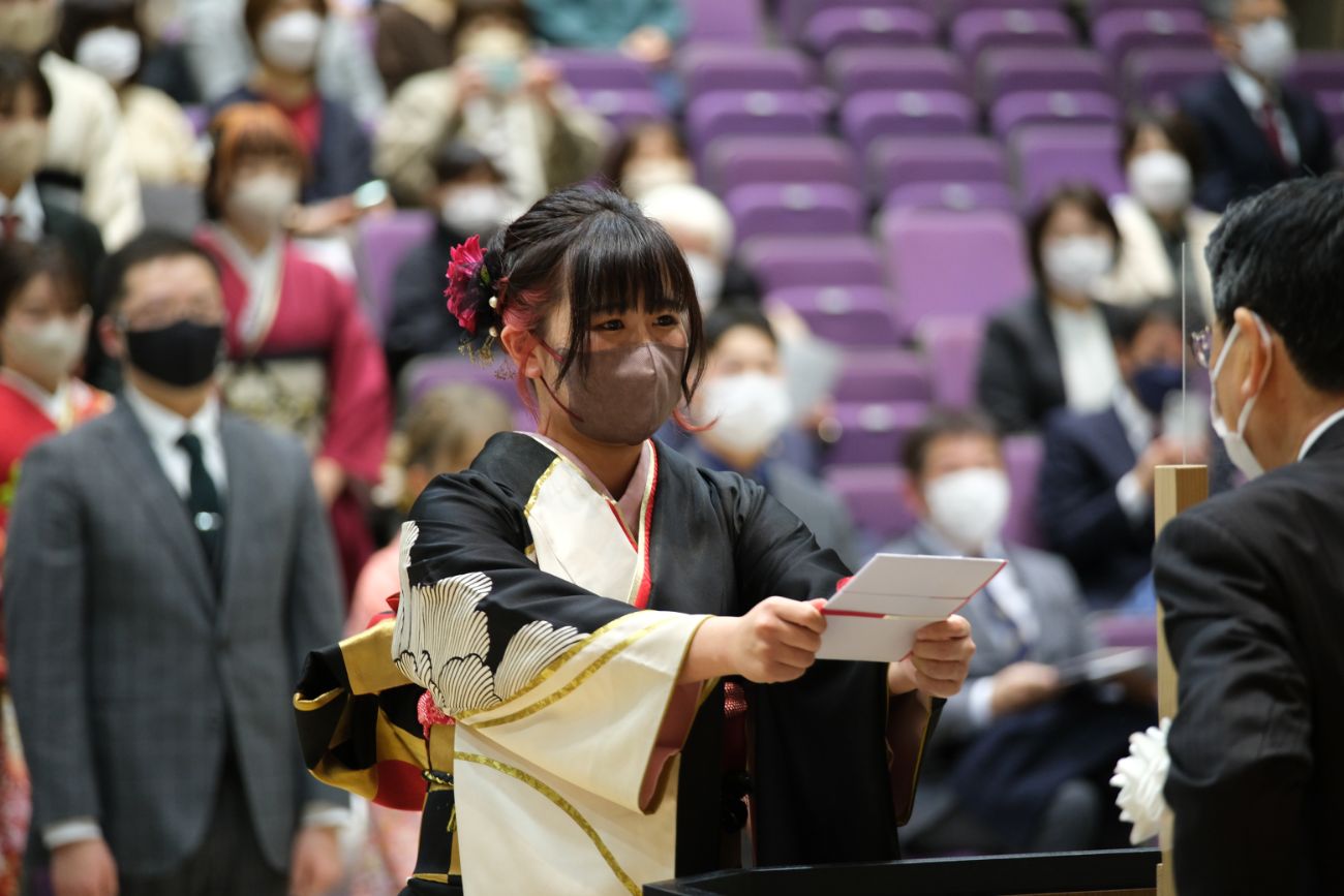 黒地に白と金の模様の振袖を着た女性が、壇上で封筒を差し出している様子の写真