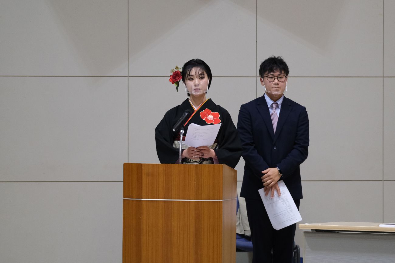 黒い振袖を着た女性が壇上でスピーチを行っており、隣には男性が立っている写真