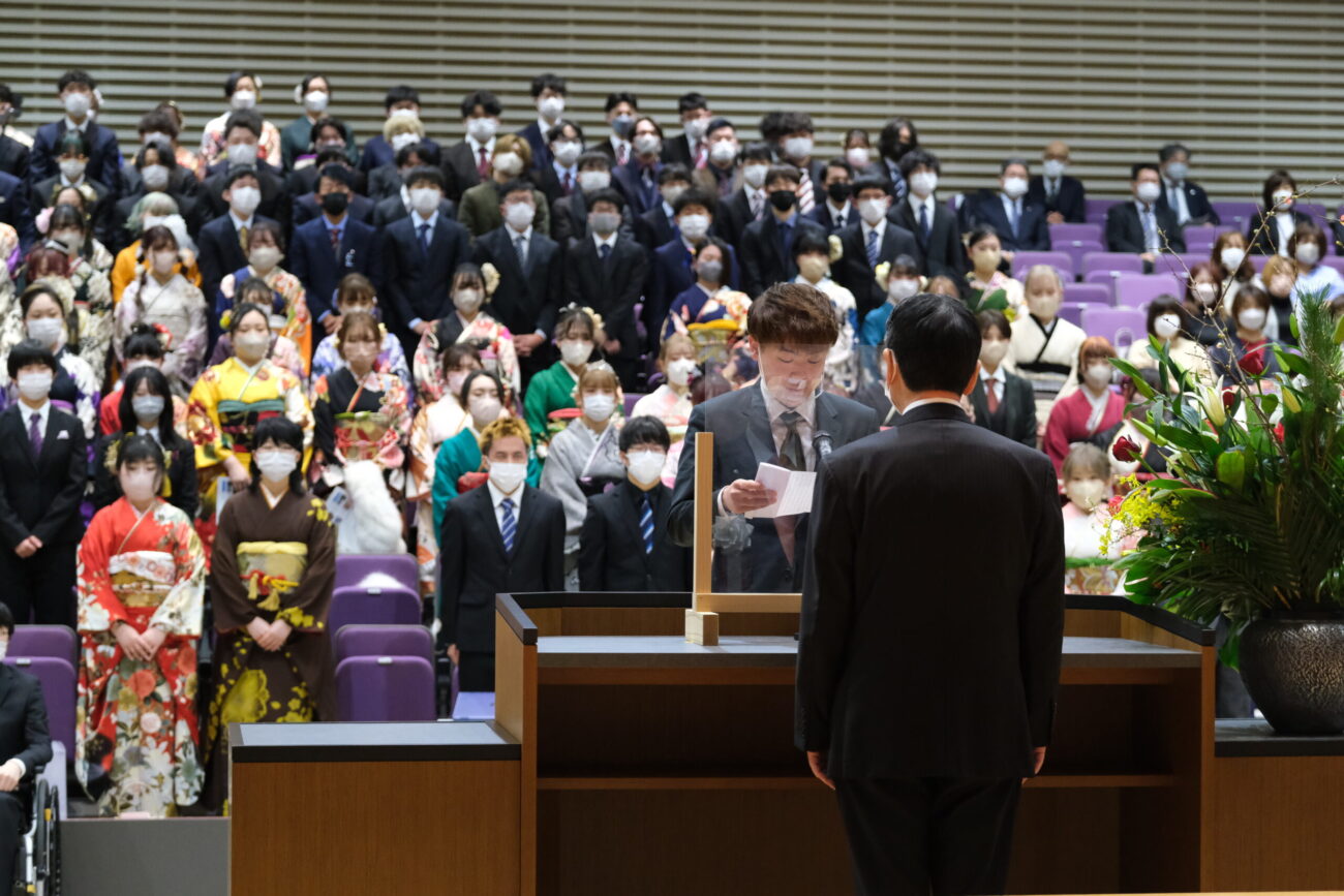 客席に座る新成人たちが立ち上がり、壇上の演台で代表者が手紙を読み上げている様子の写真