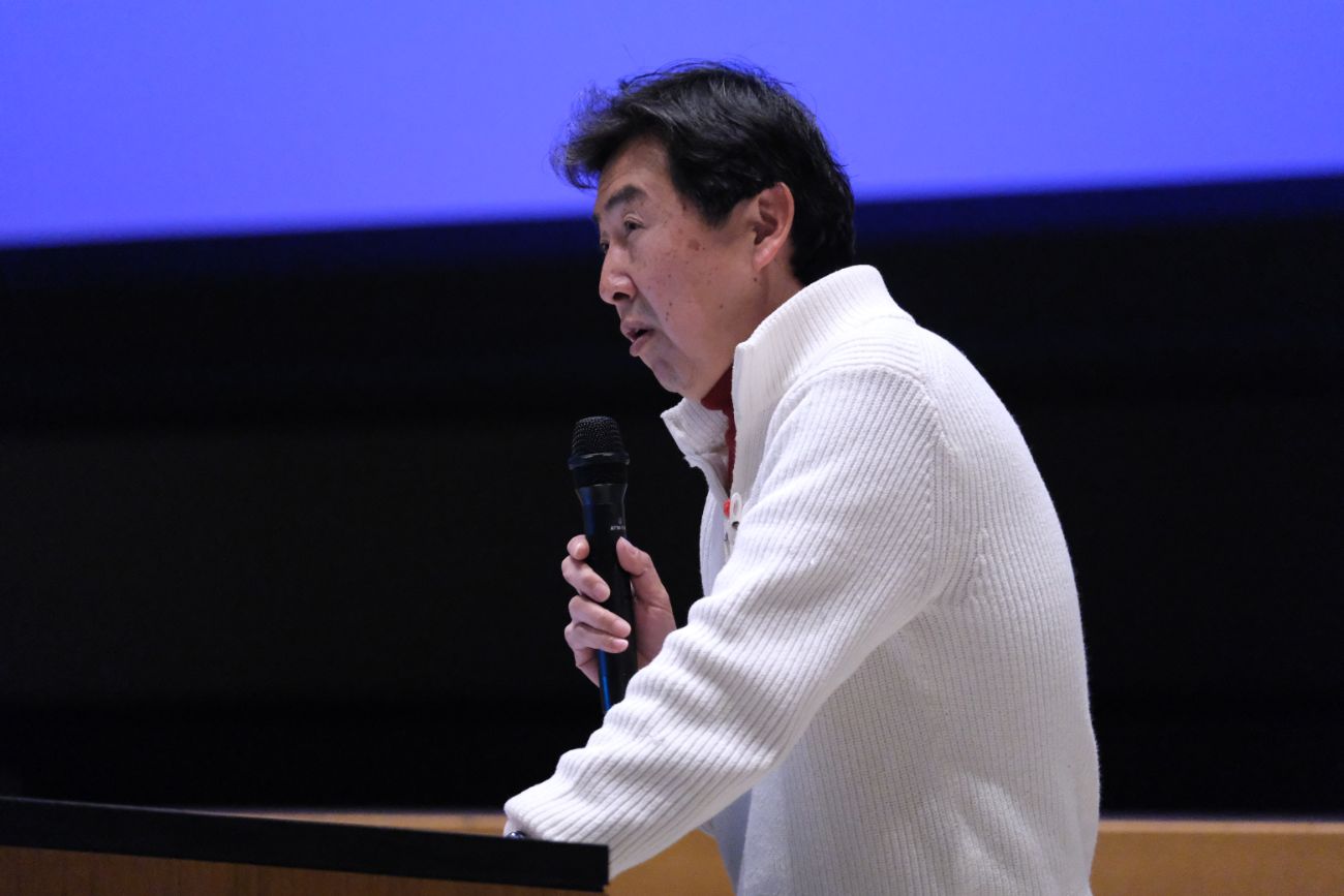 白い服装をした笠井信輔さんがマイクを持ち、壇上でスピーチをしている写真