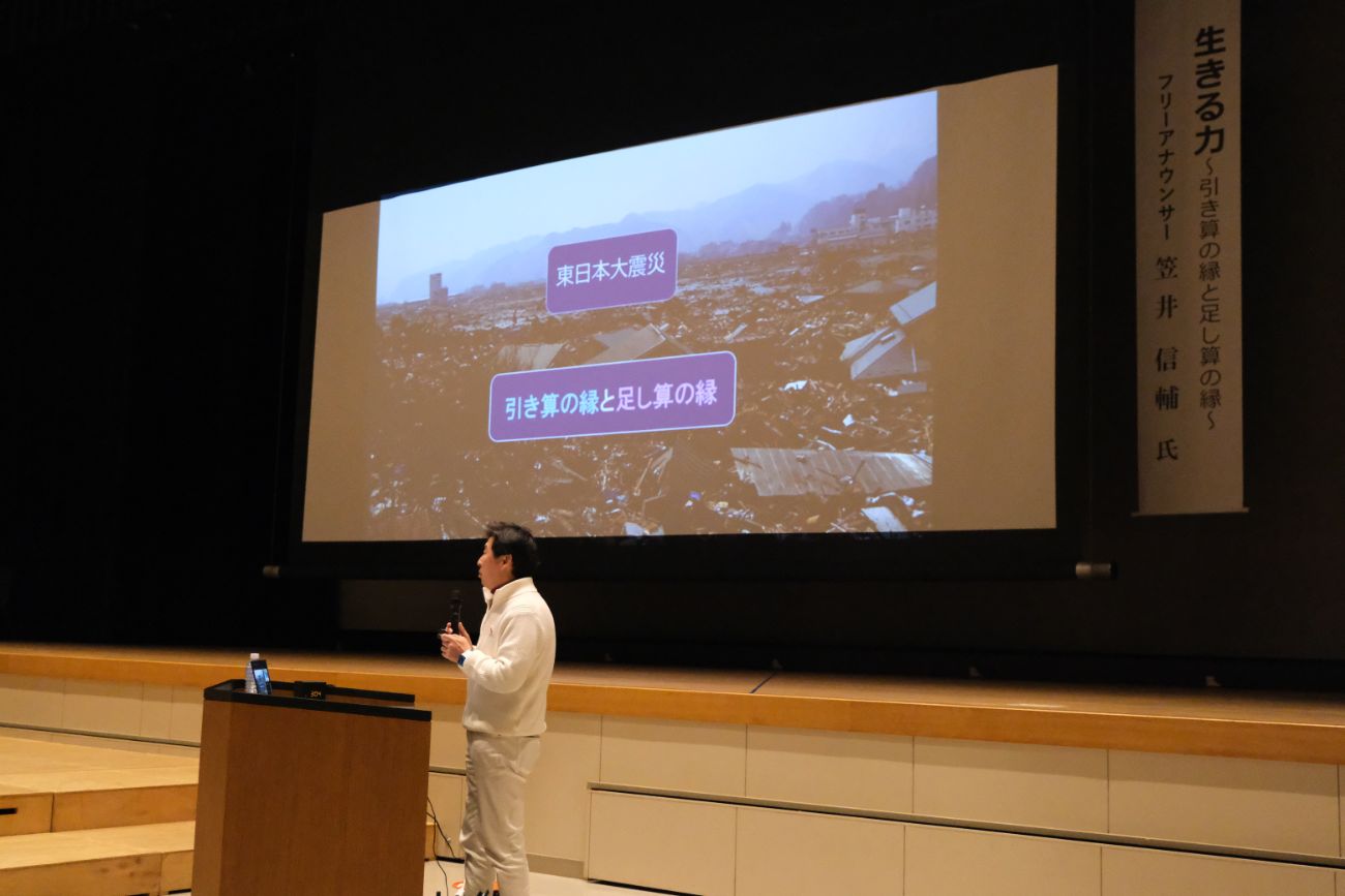 笠井信輔さんが「東日本大震災」と書かれた被災地の写真を背景に、マイクを手に講演をしている様子の写真