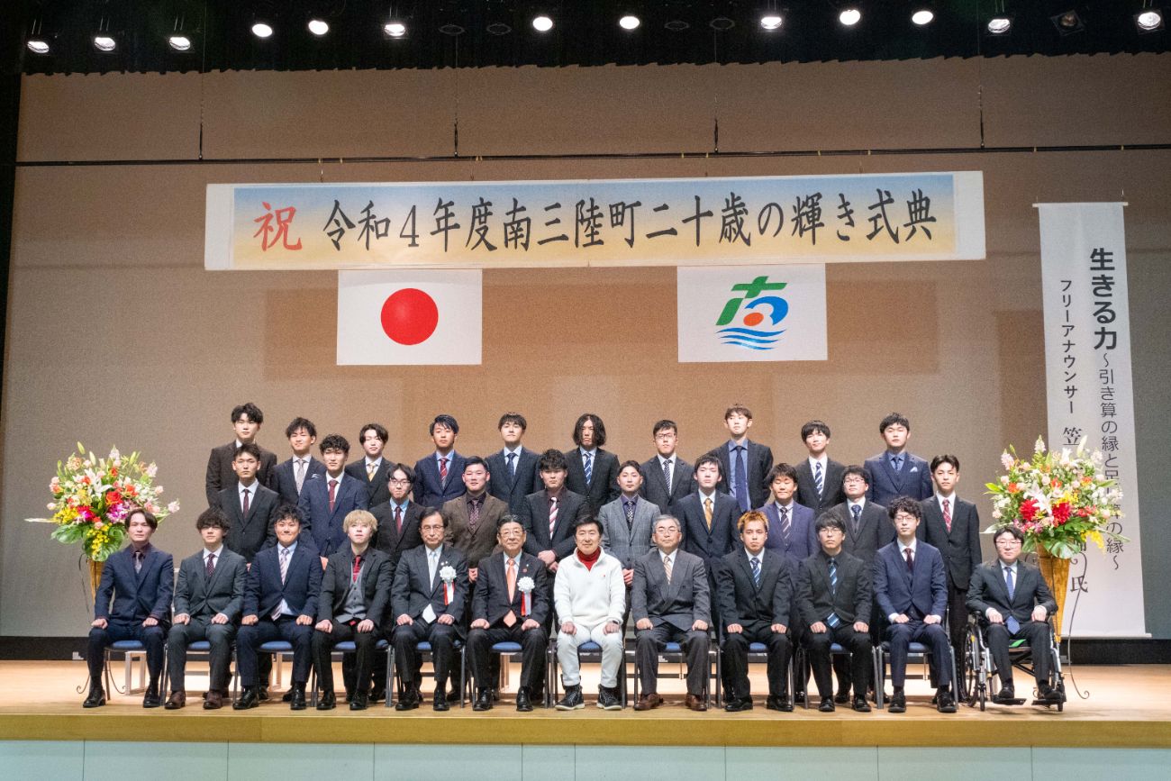 「令和4年度南三陸町二十歳の輝き式典」と書かれた横断幕が掲げられ、スーツ姿の男性ばかりが並ぶ成人式の集合写真