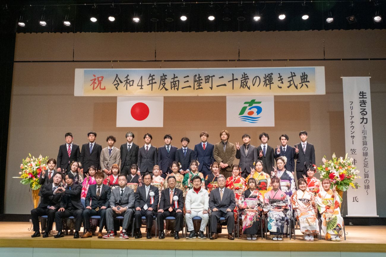 「令和4年度南三陸町二十歳の輝き式典」と書かれた横断幕が掲げられ、スーツや振袖姿の新成人たちが、壇上で記念撮影をしている集合写真
