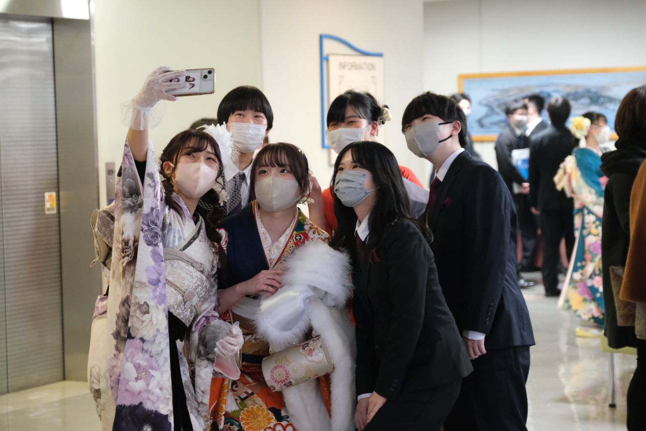 振袖やスーツを着た成人者が集まり、笑顔でスマートフォンに向かって記念撮影している様子の写真