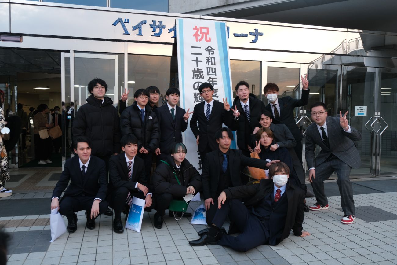 二十歳のつどいの看板の前で、スーツ姿の若者たちがピースサインをしている集合写真