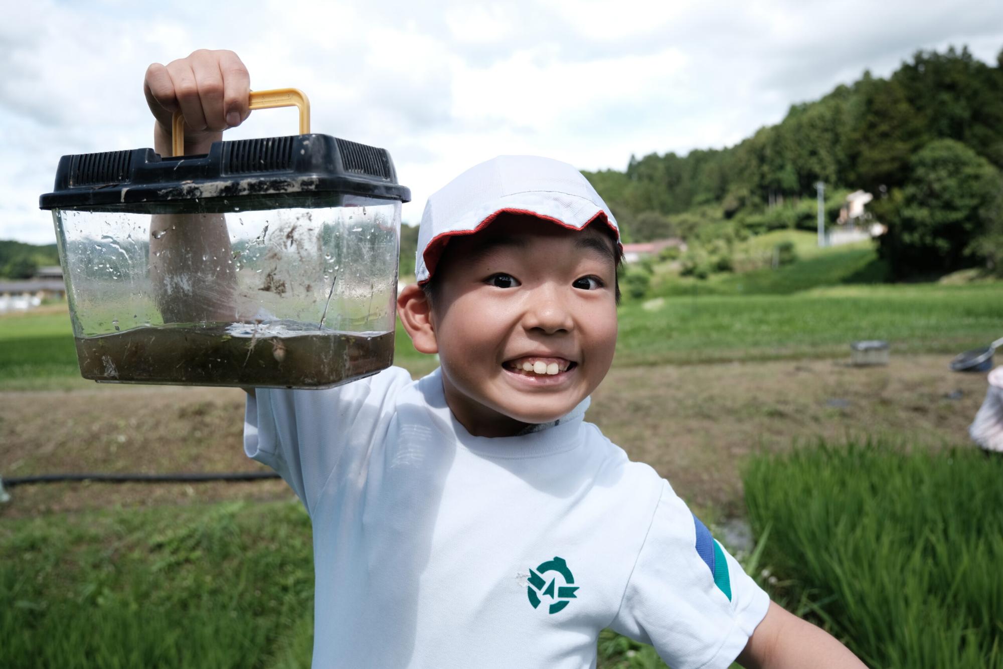 白い帽子と体操服を着た男の子が、田んぼの中で捕まえた生き物の入った透明な飼育ケースを高く持ち上げて、満面の笑みを浮かべている写真