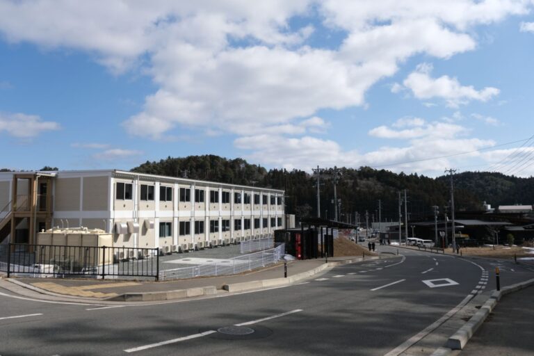 明るい色の外壁と多数の窓を備えた現代的な多層建築が、カーブした車道沿いに建っている2階建ての寮の写真