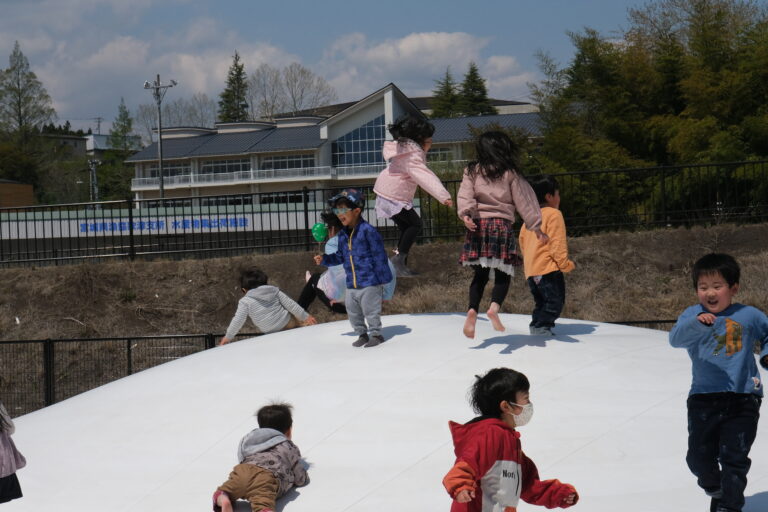 青空の下、白い「ふわふわドーム」の屋外遊具の上で、多くの子どもたちが元気に跳ねたり走ったりして遊んでいる様子の写真