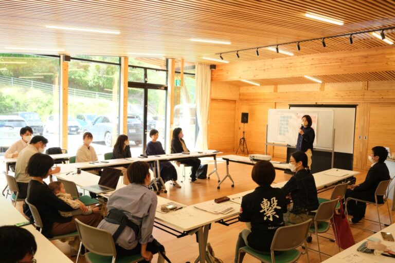 自然光が入る木造の明るい室内で、赤ちゃんを抱いた母親や地域住民らがテーブルを囲んで座り、一人の女性がホワイトボードを使って説明をしている様子の写真