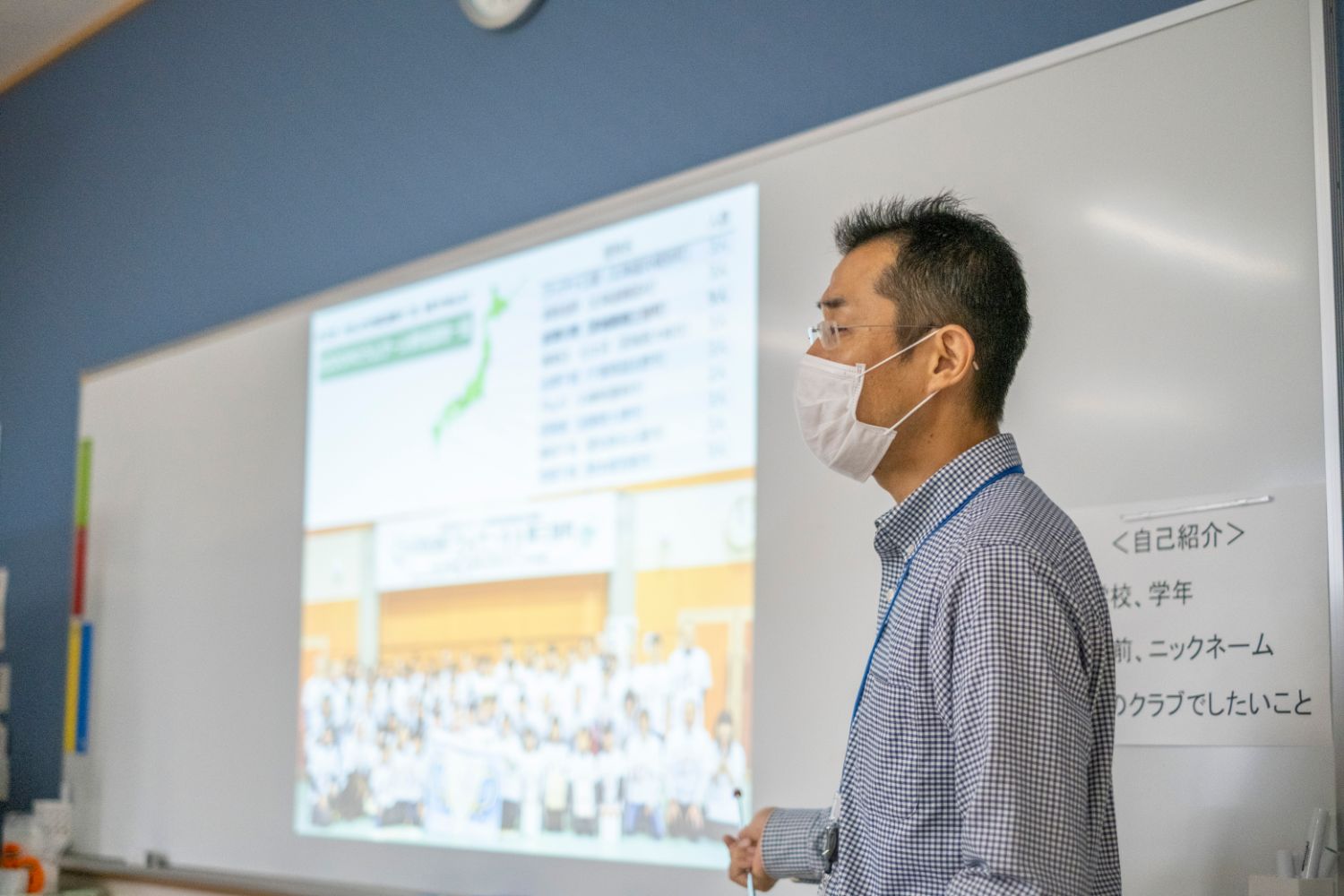 白いマスクを着けた男性が教室のホワイトボードに映し出された資料を指しながら話をしている写真