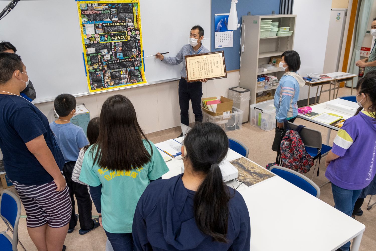 室内で複数の子どもたちが壁新聞の前に立ち、男性が指し棒で壁新聞を指し左手に表彰状を手に持っている写真