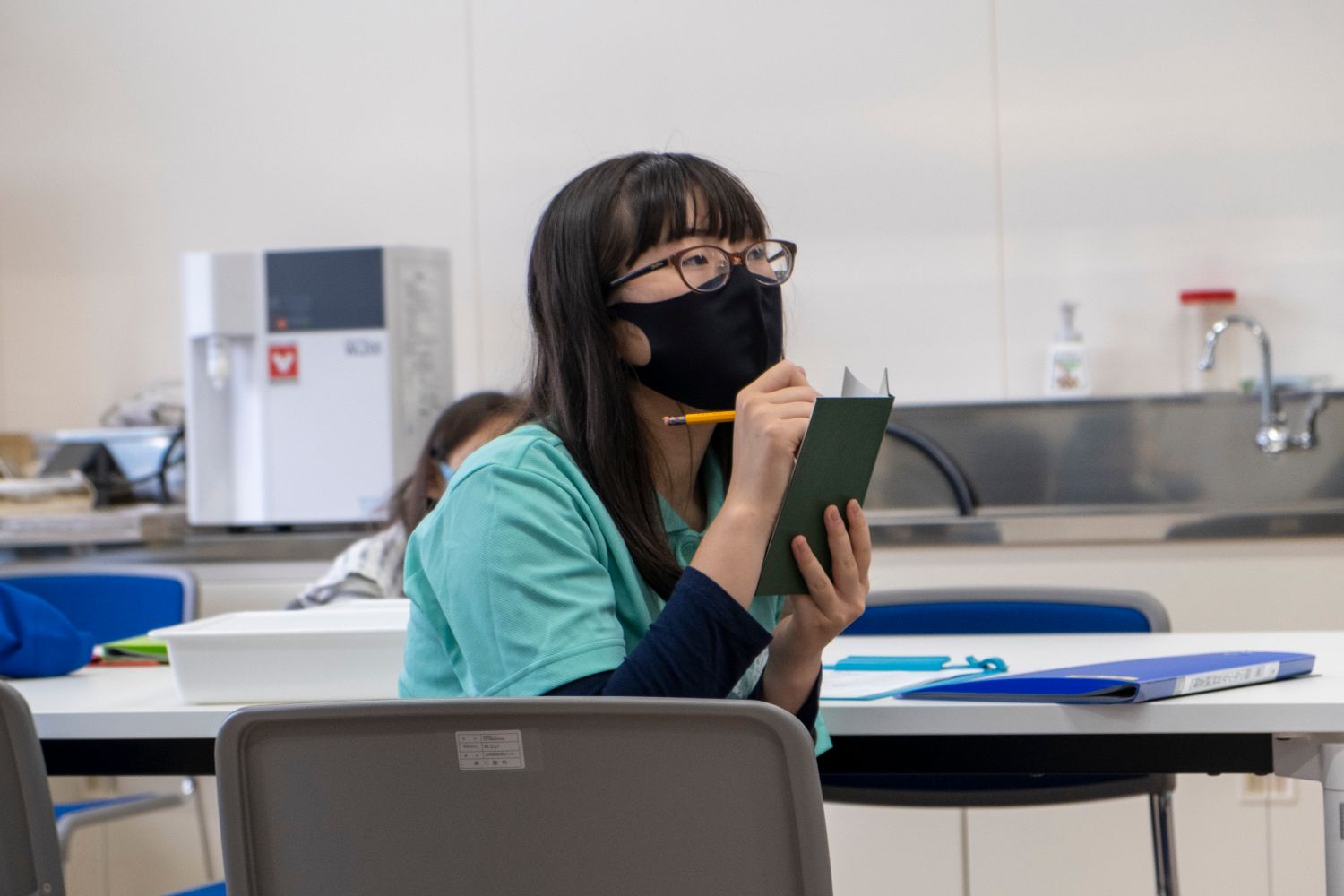室内で黒いマスクをつけ眼鏡をかけたの少女が、手に持った緑色の調査ノートに鉛筆で書きこんでいる写真
