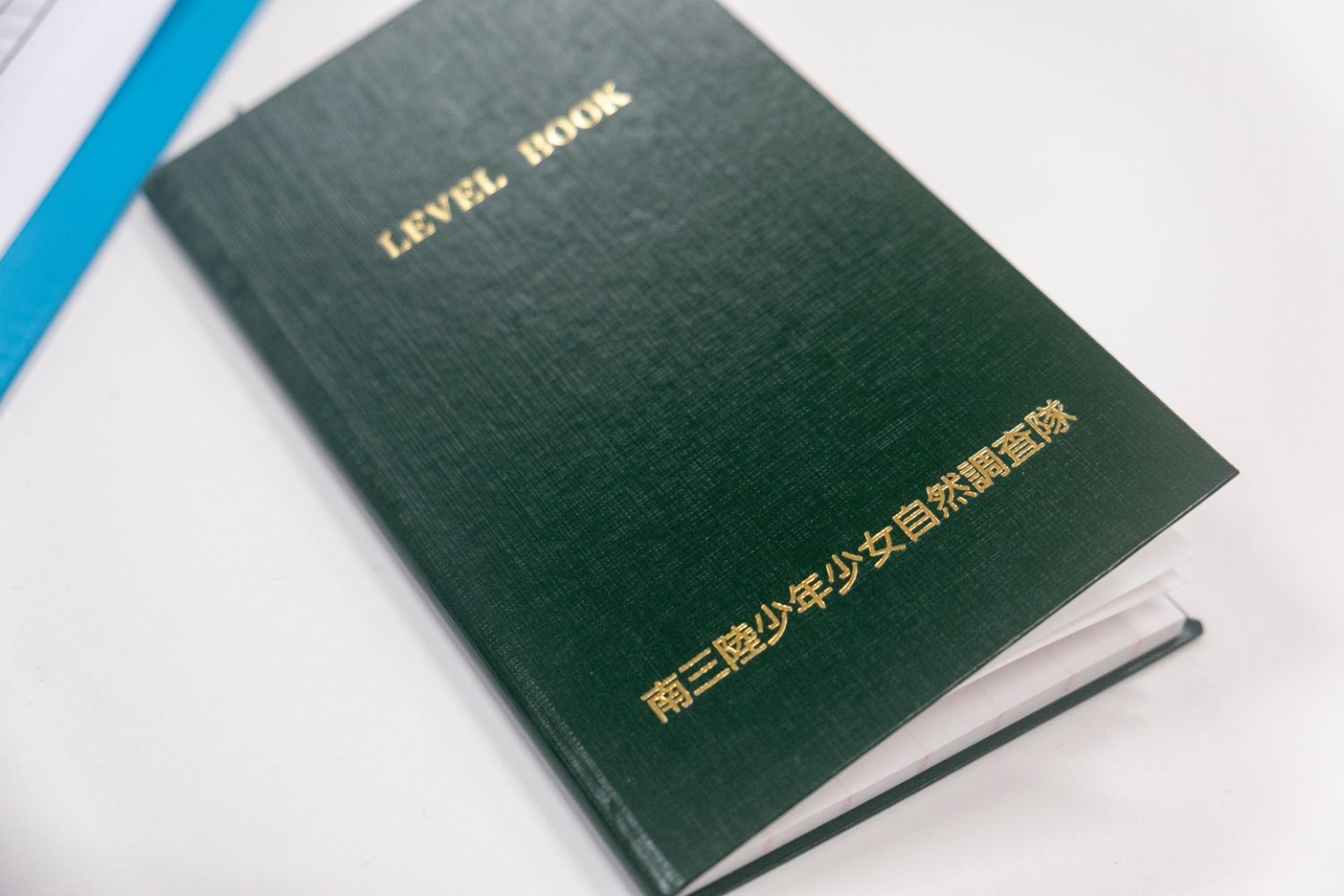 「LEVEL BOOK」と英語で書かれ、下部に日本語で「南三陸少年少女自然調査隊」と金文字で印字された緑色の表紙の記録ノートが机の上に置かれている写真