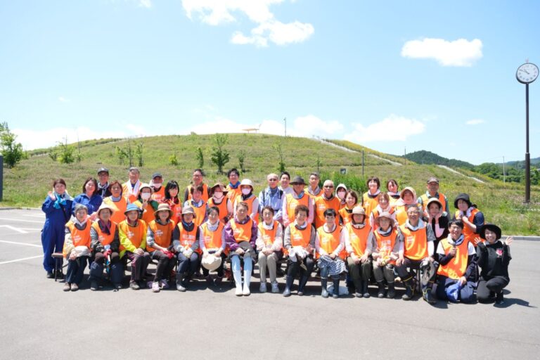 青空の下、丘のふもとの広場でオレンジ色のビブスを着た大勢の住民が整列して笑顔で記念撮影している写真