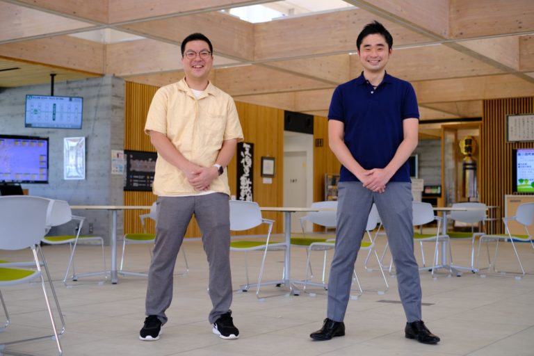 テーブルとイスが並び、大型モニターが設置された室内で、薄い黄色のシャツと青いシャツを着た2人の男性が、肩幅に両足を開き、両手を前に置いて並んで立っている写真