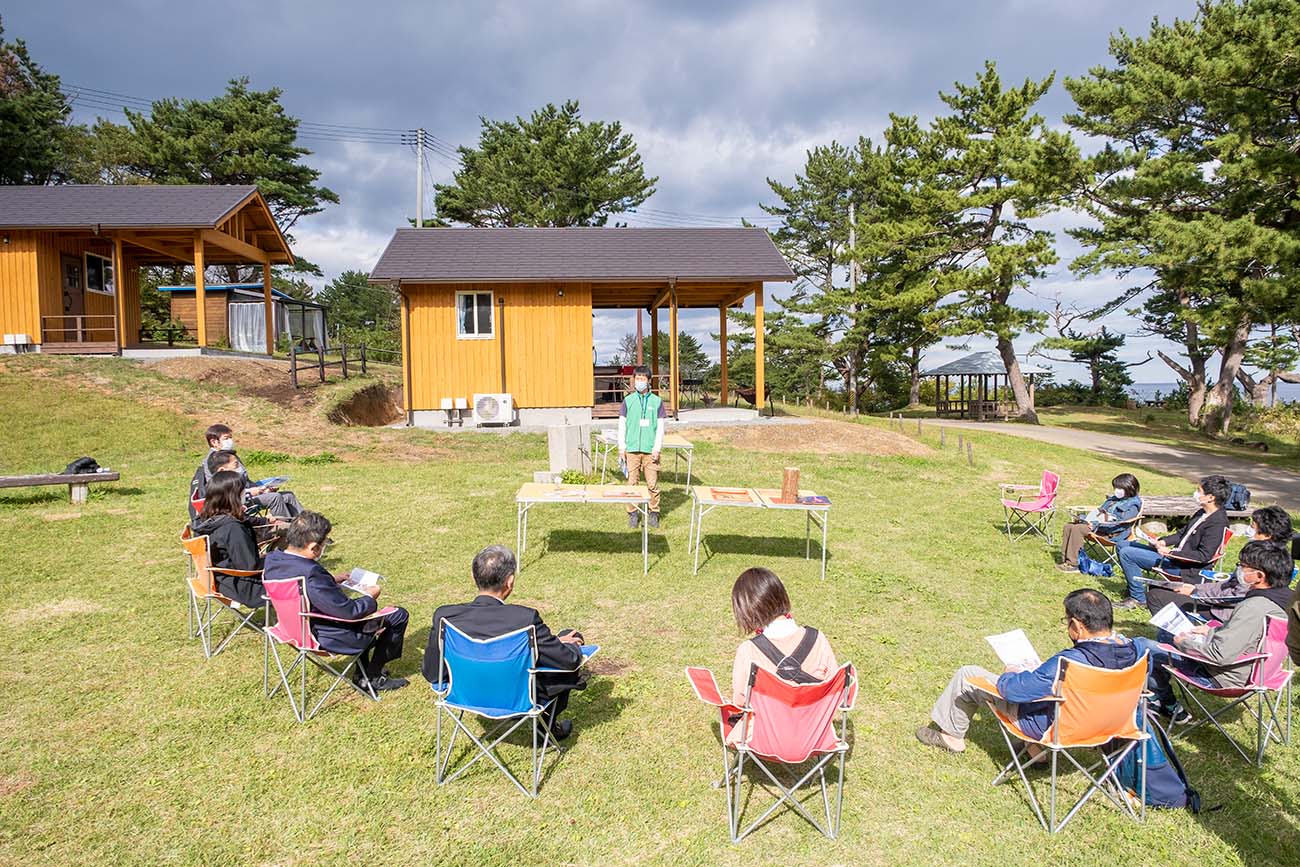 神割崎キャンプ場の新施設お披露目会に参加した地域住民や協力企業の人たちが、屋外の芝生の上で円形に座り説明を聞いている様子の写真