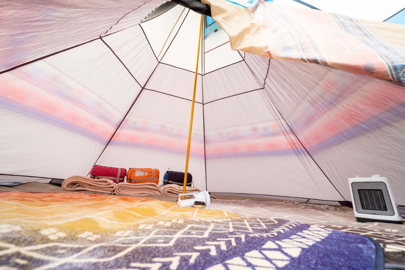 テントの中に敷かれたカラフルなラグの上に折りたたまれた毛布と収納袋が並び、テントの中央の支柱のそばに懐中電灯、右側に小型ヒーターが置かれている写真