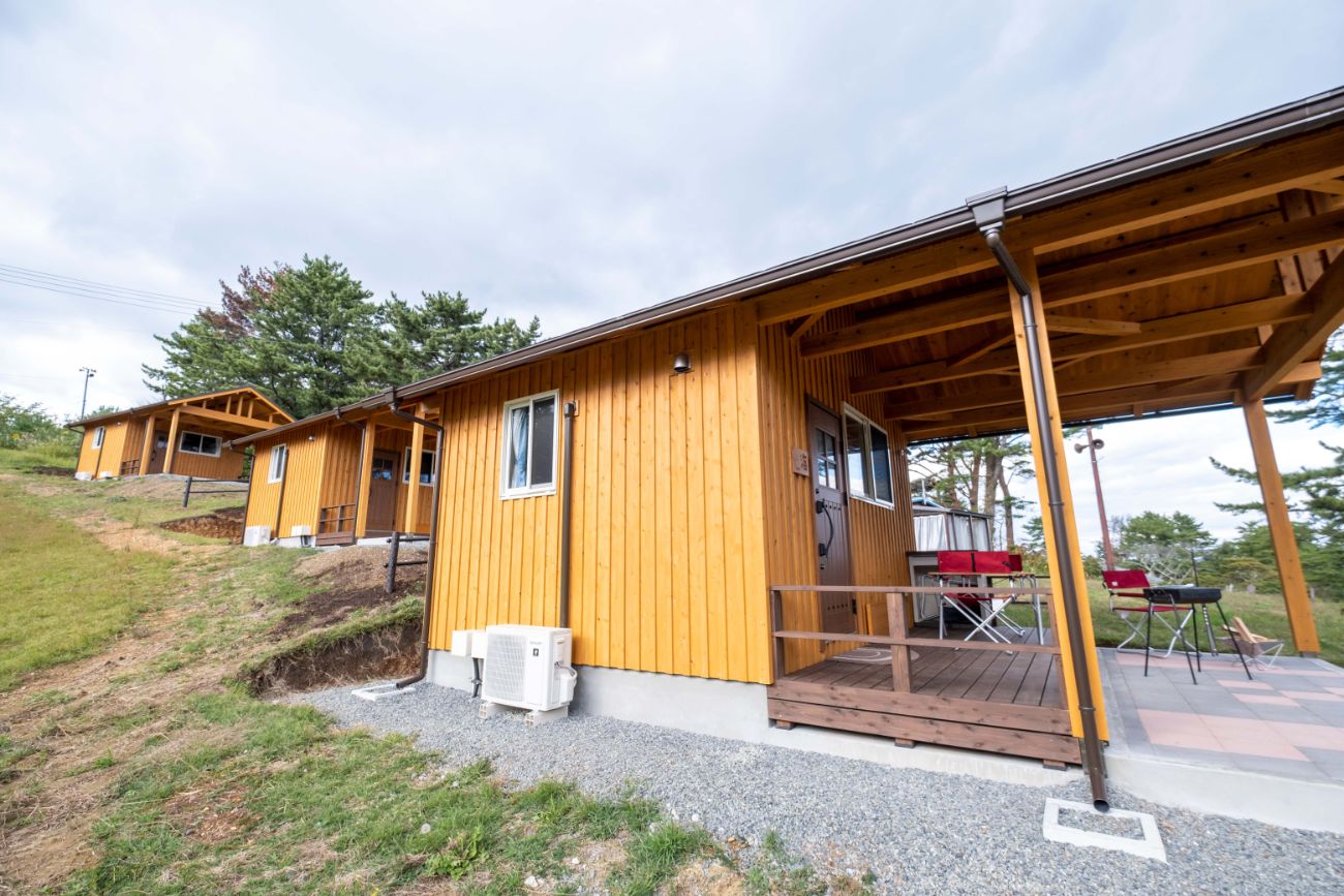 青空の下、木造のログキャビンが斜め前方から撮影されていて、テラスに赤い椅子が置かれ、右手にはハンモックが見える新ログキャビンの写真