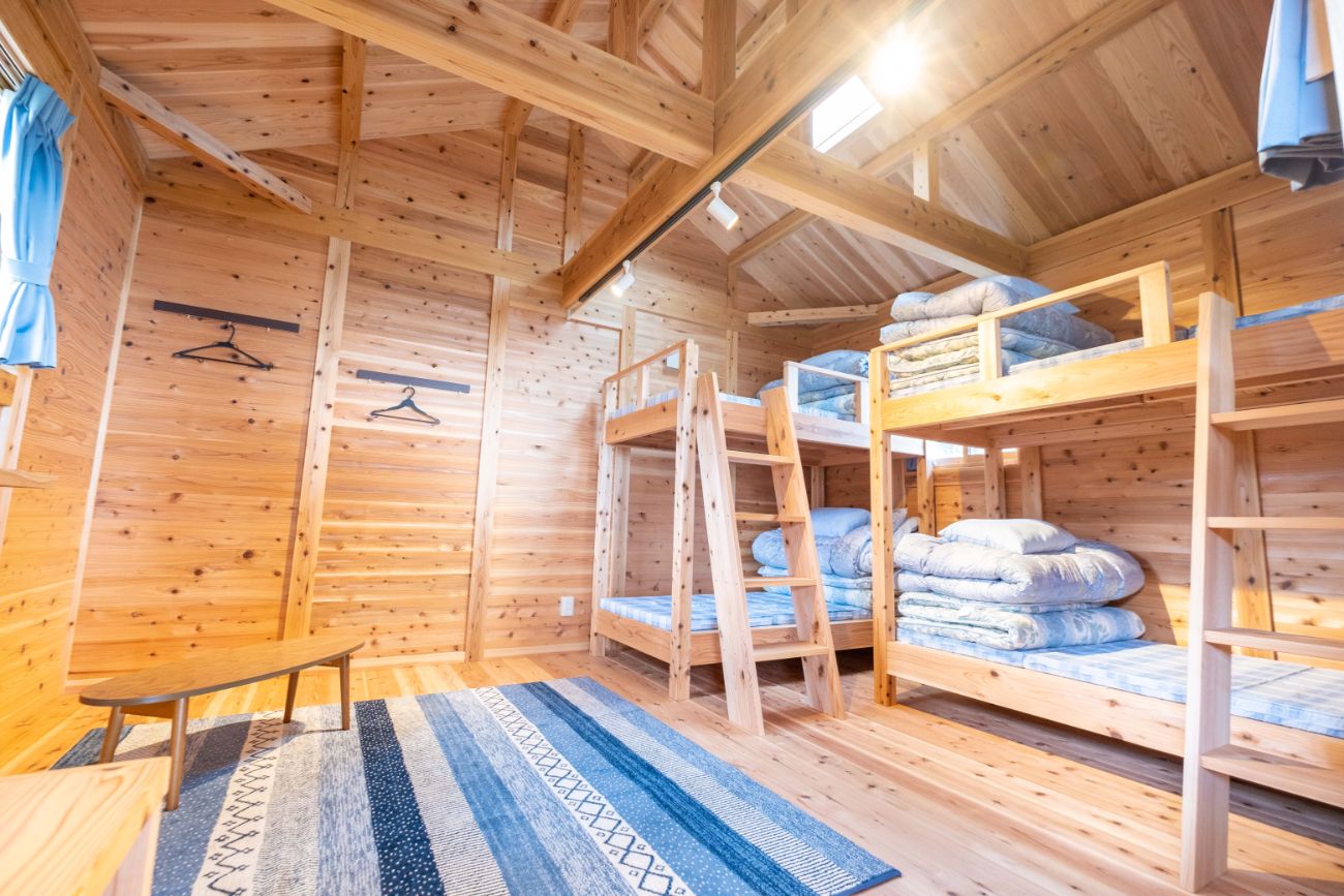 木製の壁と天井に囲まれた部屋に二段ベッドが二つあり、それぞれのベッドに布団や枕が積まれ、床には青と白のストライプ模様のラグが敷かれて、テーブルが置かれているログキャビンの室内の写真