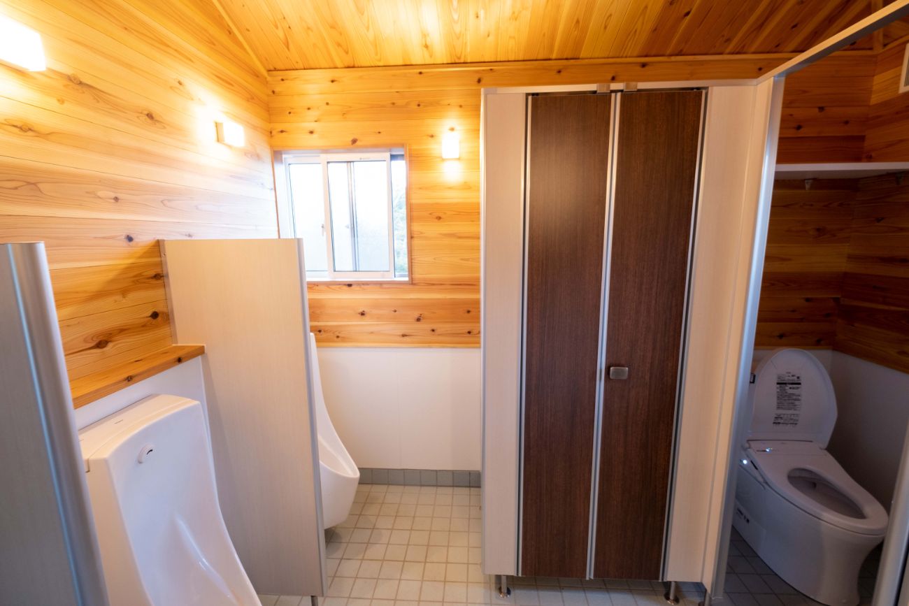 木目調の壁と明るい照明がある男女別の仕切り付きトイレの中で、左側に白い小便器が2台設置され、右側にドアのついた個室トイレが見える室内の写真